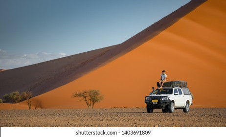 Young Asian man tourist sitting on camper car near orange sand dune landscape. Travel desert in Namibia, Africa.
