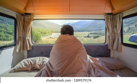 Young Asian Man Traveler Staying In The Blanket Looking At Mountain Scenery Through The Window In Camper Van In The Morning. Road Trip In Summer Of South Island, New Zealand.