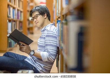 Young Asian Man University Student Reading Book Sitting By Bookshelf In College Library For Education Research And Self Improvement. Scholarship And Educational Opportunity. World Book Day Concept