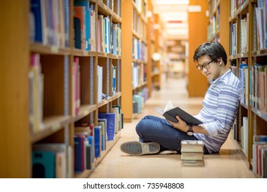 Young Asian Man University Student Reading Book Sitting By Bookshelf In College Library For Education Research And Self Improvement. Scholarship And Educational Opportunity. World Book Day Concept