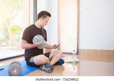 Young Asian Man Using Mobile Phone While Lifting Dumbell At Home