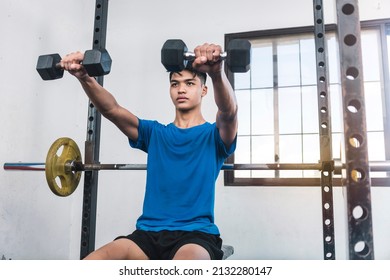 A young asian man wearing a blue shirt and seated on a bench does dumbbell front raises. Shoulder and upper body workout at the gym. - Powered by Shutterstock