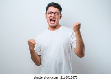 Young asian man wearing casual shirt standing over isolated white background very happy and excited doing winner gesture with arms raised, smiling and screaming for success. Celebration concept