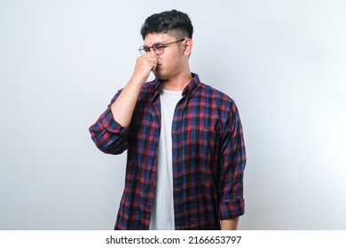 Young asian man wearing casual shirt over white background smelling something stinky and disgusting, intolerable smell, holding breath with fingers on nose. Bad smell