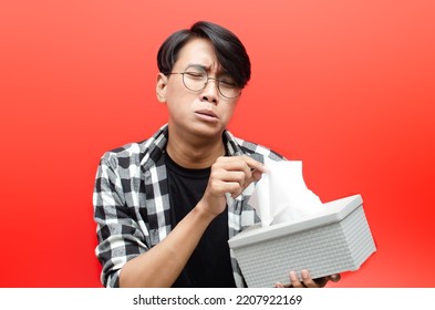 Young Asian Man Wearing Glasses With Crying Face Expression Holding Tissue Box Isolated On Red Background. Crying Face Expression Of Young Asian Man With Hand Gesture. 