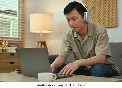 Young Asian man wearing headphone sitting on couch working remotely online on laptop at home - Shutterstock ID 2310732907