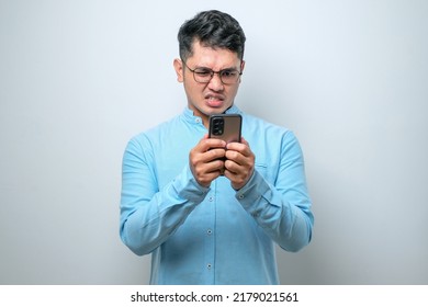 Young Asian Man Wearing Plain Shirt Standing Over White Background Feels Angry Looking At Smartphone