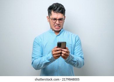 Young Asian Man Wearing Plain Shirt Standing Over White Background Feels Angry Looking At Smartphone