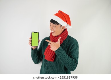 Young asian man wearing santa clause hat, red scarf and green long sleeved sweater is showing smartphone green screen, isolated over white background. - Powered by Shutterstock