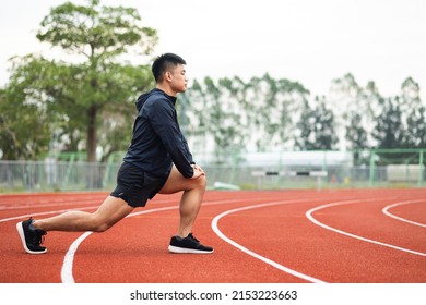 Young Asian Man Wearing Sportswear Warming Up Front Of Sport Stadium. Chinese Man Stretching Legs Before Running On Track Exercise In The Morning. Healthy And Athlete Lifestyle Concept.