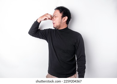 Young Asian man wearing t-shirt standing over isolated white background smelling something stinky and disgusting, intolerable smell, holding breath with fingers on nose. Bad smells concept. - Shutterstock ID 2259204367