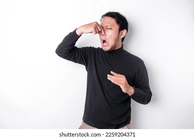 Young Asian man wearing t-shirt standing over isolated white background smelling something stinky and disgusting, intolerable smell, holding breath with fingers on nose. Bad smells concept. - Shutterstock ID 2259204371