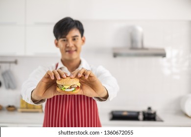 A young Asian man wearing a white shirt and red apron holding a burger in a white kitchen. - Powered by Shutterstock