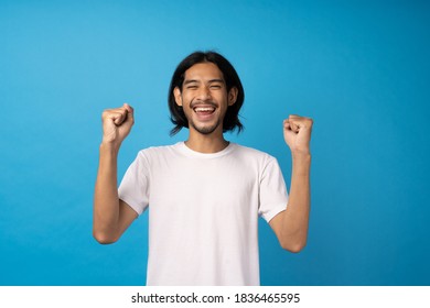 Young Asian man wearing a white T-shirt is showing signs of joy, raising his fist. Man standing with his hand raised and looking at the camera split up against a blue background.