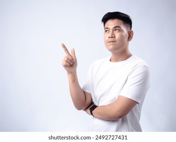 A young Asian man, wearing a white shirt and black watch, looks thoughtfully upward while pointing with his right index finger. The plain white background highlights the subject's contemplative - Powered by Shutterstock