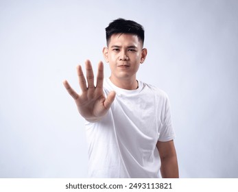 A young Asian man, wearing a white shirt, holds his hand up in a stop gesture with a serious expression. The plain white background emphasizes the subject's firm and assertive demeanor - Powered by Shutterstock