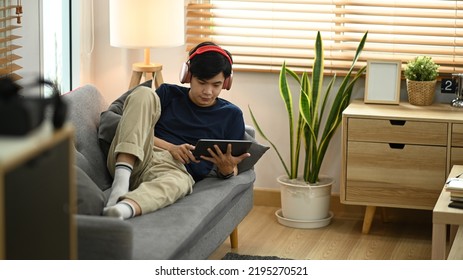 Young Asian Man Wearing Wireless Headphone Watching Online Movie Or Browsing Internet On Digital Tablet