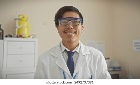 A young asian man in a white lab coat and protective eyewear smiles in a hospital setting, exuding professionalism and approachability. - Powered by Shutterstock