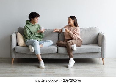 Young Asian Man And Woman Drinking Coffee And Talking While Sitting On Couch At Home. Loving Millennial Couple Enjoying Coffee While Having Conversation, Spending Weekend Together