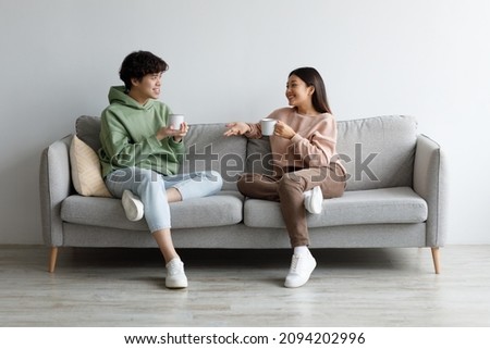 Young Asian man and woman drinking coffee and talking while sitting on couch at home. Loving millennial couple enjoying coffee while having conversation, spending weekend together