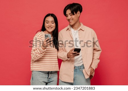 Young asian man and woman wearing casual clothing using mobile phones while standing isolated over red background