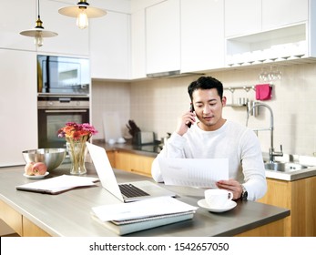 Young Asian Man Working From Home Sitting At Kitchen Counter Looking At A Document And Talking On Cellphone