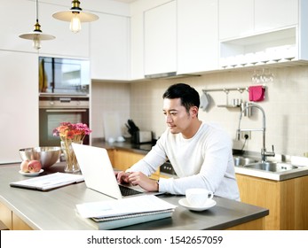 Young Asian Man Working From Home Sitting At Kitchen Counter Using Laptop Computer