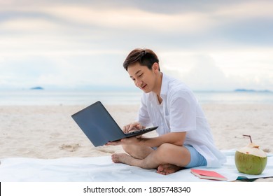 Young Asian Man Working Outdoor During His Vacation Time By Laptop While Sitting On The Beautiful Beach. Summer, Holidays, Vacation And Happy People In Thailand Concept.
