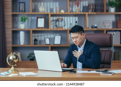 Young Asian man in the workplace is experiencing chest pain, heart attack, stroke. Holds on to his chest, difficulty breathing, wincing in pain. In the office, sitting at a table - Shutterstock ID 2144928487