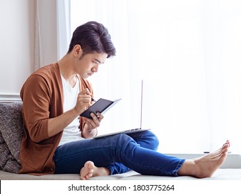 Young Asian Man Writing In A Book When Using Laptop