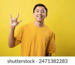 A young Asian man in a yellow shirt smiles and makes a hand sign for "I love you" in American Sign Language against a matching yellow background.
