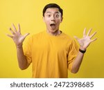 Young Asian man in a yellow shirt against a yellow background, with a surprised and excited expression, hands raised, and mouth open wide. Ideal for themes related to surprise