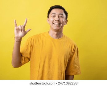 A young Asian man in a yellow shirt smiles and makes a hand sign for "I love you" in American Sign Language against a matching yellow background. - Powered by Shutterstock