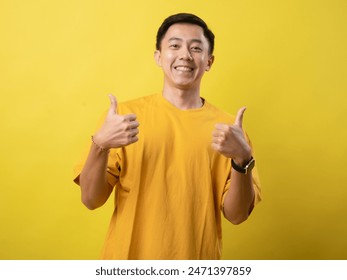 A young Asian man in a yellow shirt gives a thumbs up and smiles confidently against a solid yellow background. This image portrays positivity, approval, and satisfaction - Powered by Shutterstock