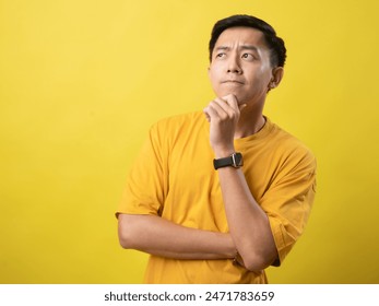 A young Asian man in a yellow shirt stands thoughtfully with his hand on his chin and arms crossed, against a solid yellow background - Powered by Shutterstock