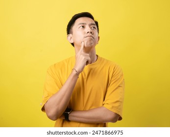 A young Asian man in a yellow shirt stands thoughtfully with his hand on his chin and arms crossed, against a solid yellow background - Powered by Shutterstock