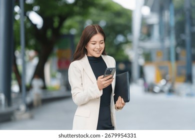 Young Asian manager using mobile phone and holding a document folder while walking near office buildings