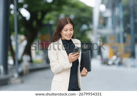Young Asian manager using mobile phone and holding a document folder while walking near office buildings