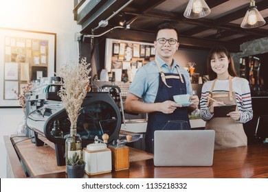 Young Asian Man&woman Barista Wear Apron Using Laptop With Coffee Cup Served To Customer At Bar Counter In Coffee Shop With Smile Face.Concept Of Cafe And Coffee Shop Small Business.