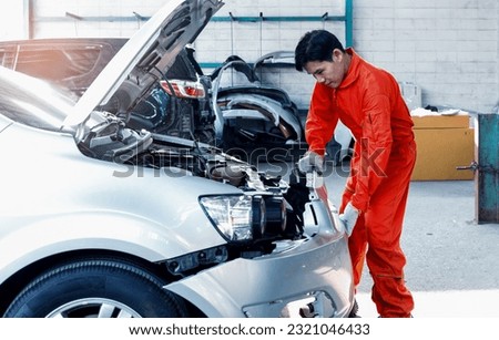 Young Asian mechanic in orange uniform changes front bumper Repair car body after road accident in a garage. Repair service. Skilled mechanic working behind the scenes with cars and car parts.