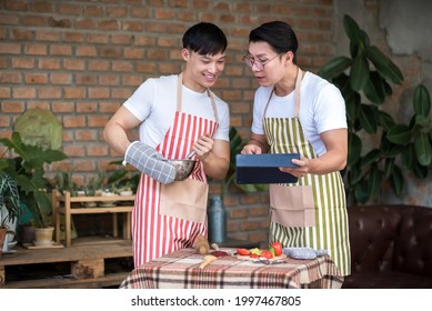 

Young Asian Men Couple Friend Using Tablet While Cooking In The Kitchen At Home For Holiday Festival Celebration.. LGBT, Gay And Love Relationship Concept