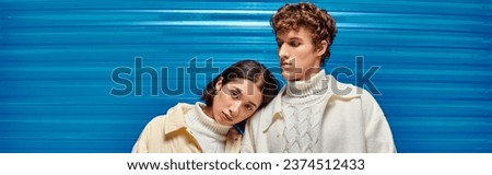 young asian model leaning on shoulder of stylish man on blue plastic backdrop, winter trend, banner