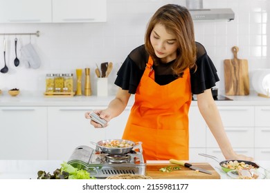 Young Asian mom wearing orange apron learn to cook for family with happy and drop peppers into food on flying pan on gas stove to enhance taste for kid in modern home kitchen