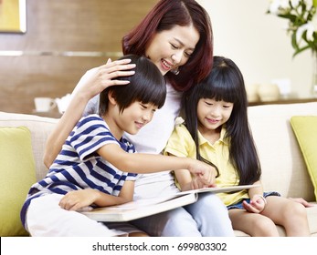 Young Asian Mother And Children Sitting On Couch At Home Reading A Book Together, Happy And Smiling.