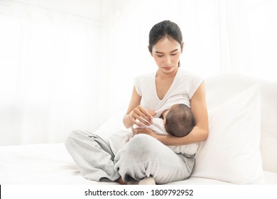 Young asian mother feeding breast her baby on bed at home in white room. Asia mom holding her baby and looking to her child.  Woman and new born relax in a white bedroom.