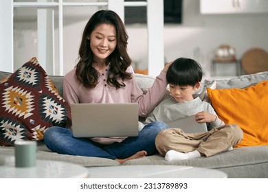 young asian mother and five-year-old son sitting on couch at home using laptop computer and digital tablet - Powered by Shutterstock