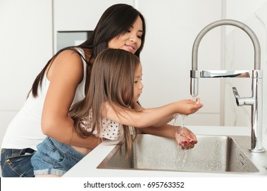 Young Asian Mother Helping Her Daughter To Wash Hands In A Sink On A Kitchen