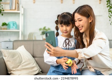 Young Asian Mother And Her Daughter Are Making Selfie Using A Phone, Hugging And Smiling While Sitting On The Sofa At Home. 