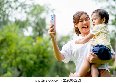 Young Asian Mother Hold Little Boy And Use Mobile Phone For Selfie Photo Shoot In Garden And They Look Happy With Holiday Time In The Morning Together.