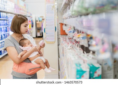 Young Asian Mother And Little Cute Baby In Carrier Are Shopping Newborn Baby Product In Supermarket. Mom Is Choosing New Nipple For Her Child.